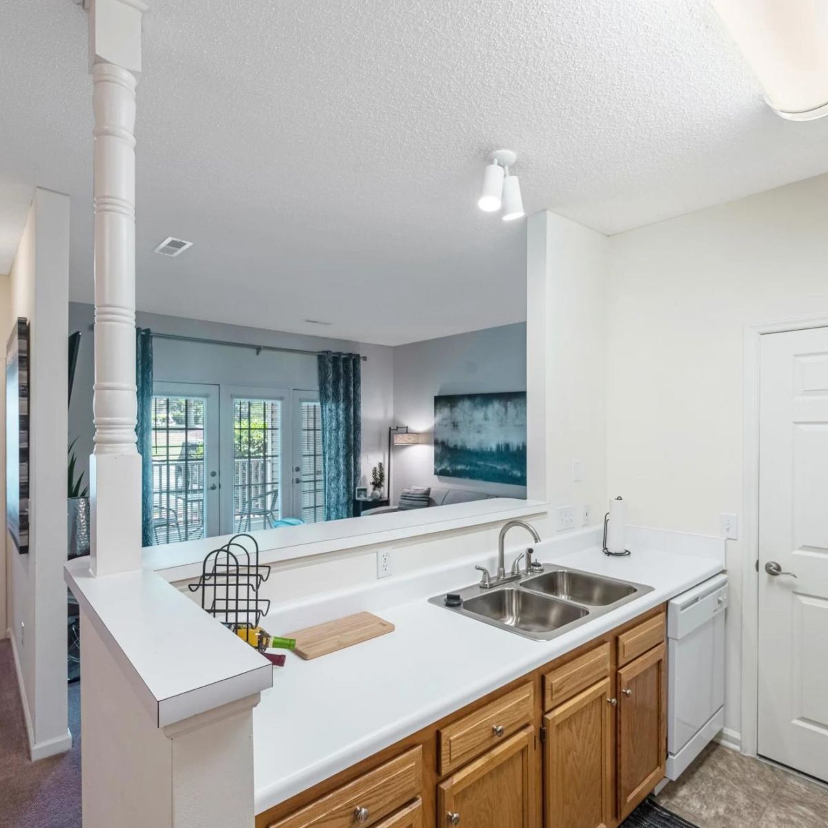 Kitchen with double sink and wood cabinets, overlooking a living area with large windows and a bedroom nearby.