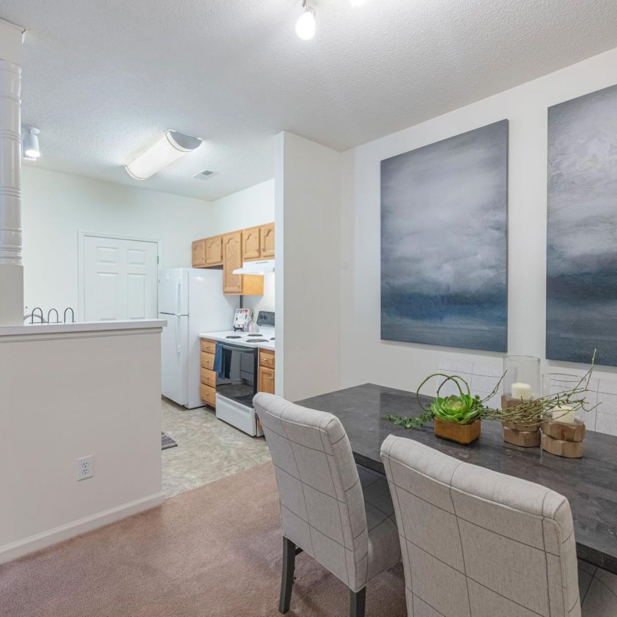 Modern dining area with gray chairs, black table, wall art, and a view into a white kitchen with wood cabinets.
